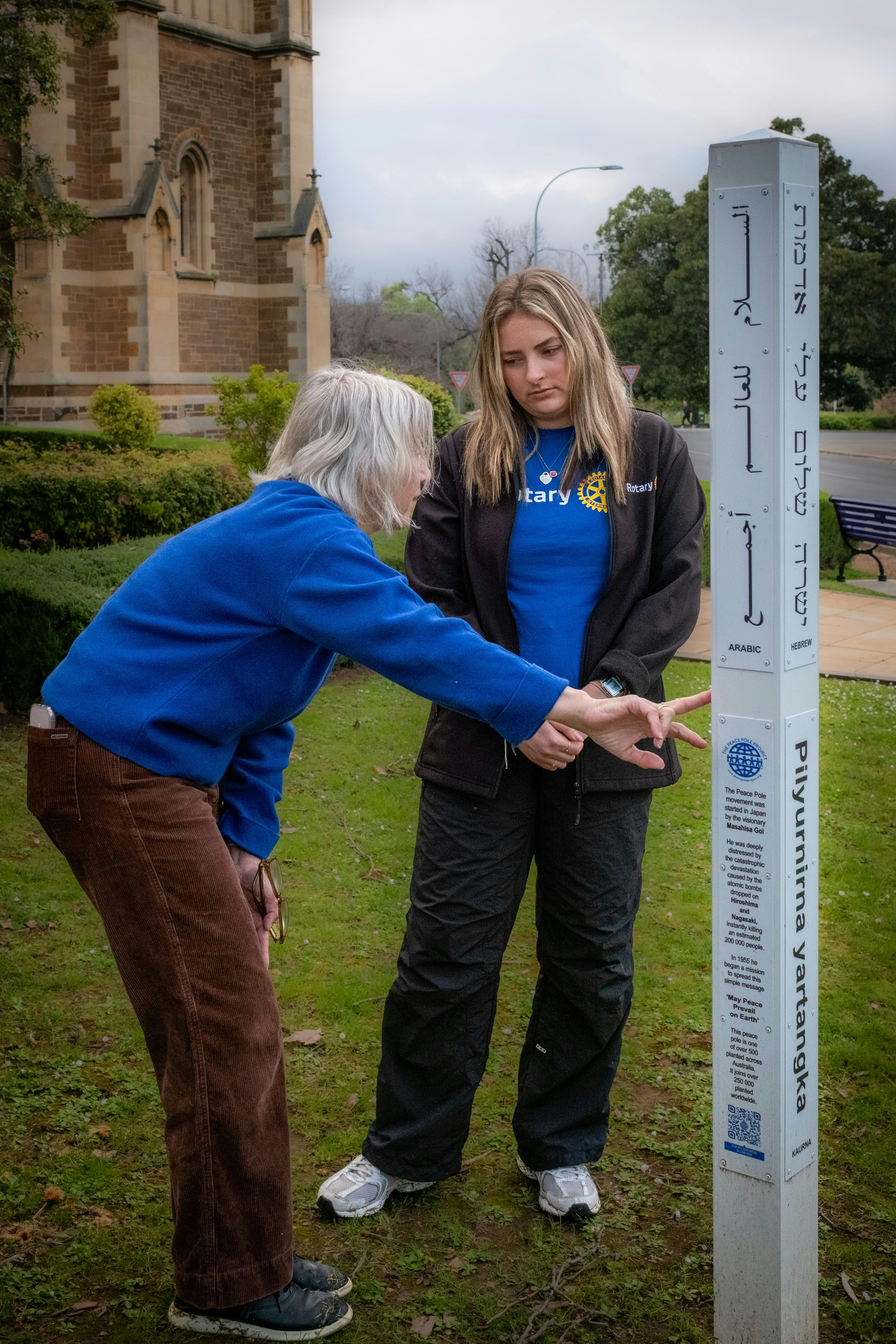 Two women look at and touching a white pole with the words May Peace Prevail on Earth in languages