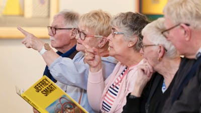 A photograph of 5 people in a gallery space pointing and looking forward
