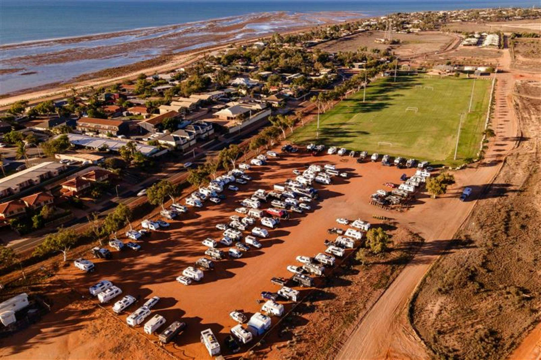 Aerial view of RV overflow with rows of caravans on red dirt beside sports field and coast