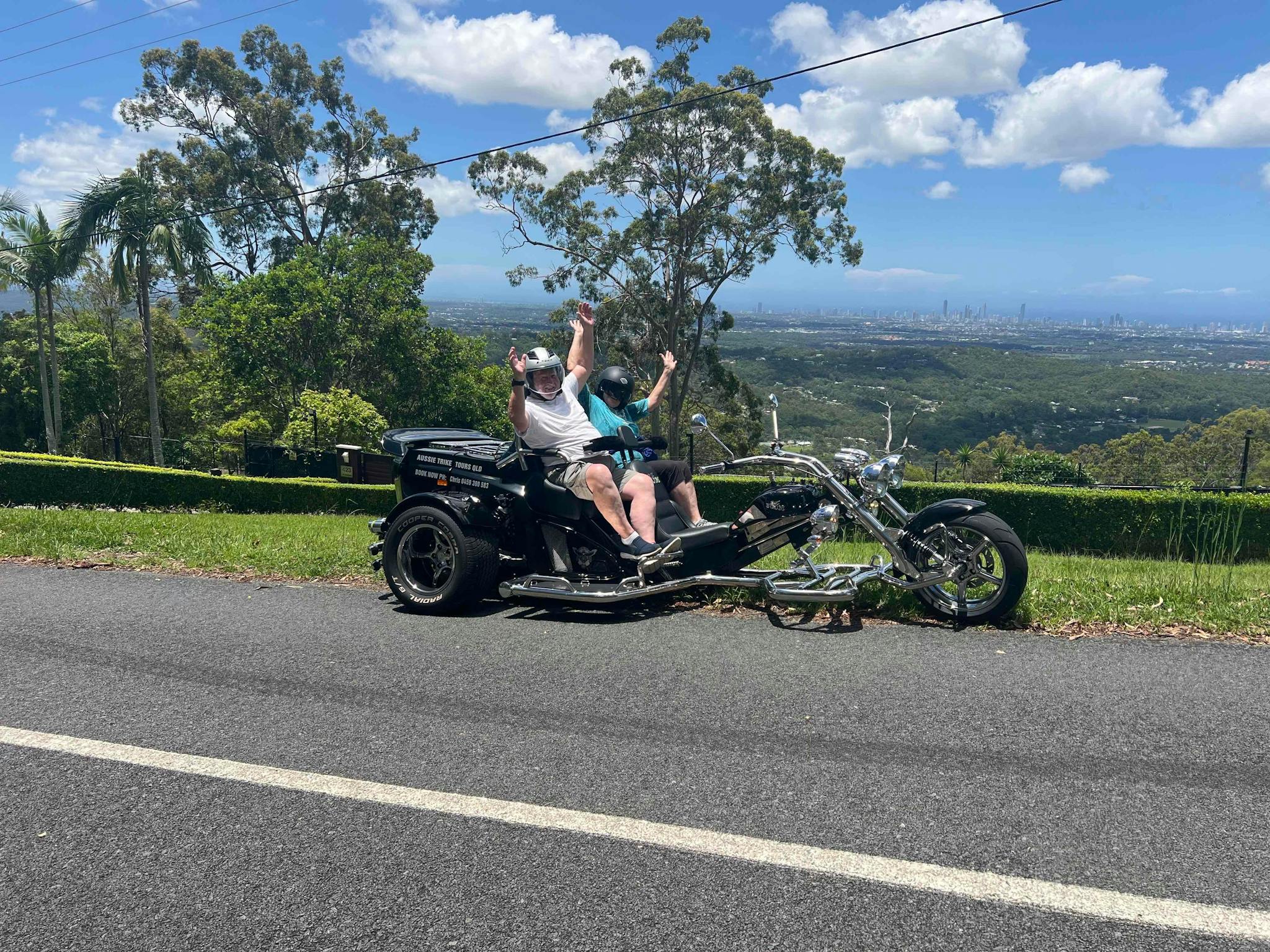 Image shows two people on the back of a trike at the top of a lookout on a sunny day. Image shows two people on the back of a trike at the top of a lookout on a sunny day.