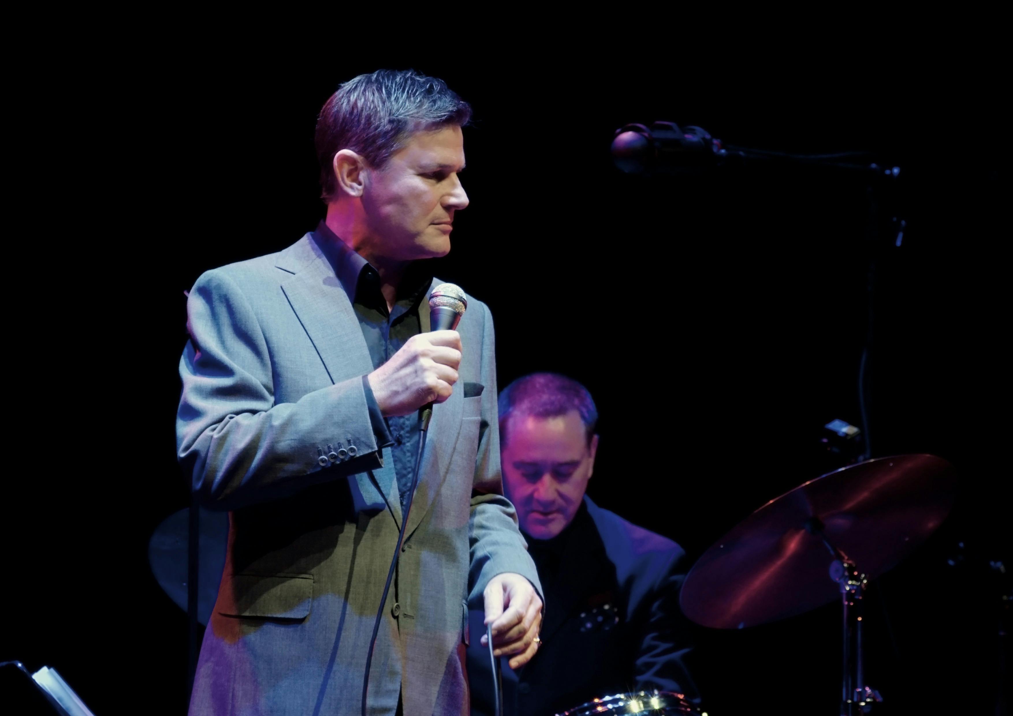 Man in grey suit holding a microphone with a man on drum kit behind him
