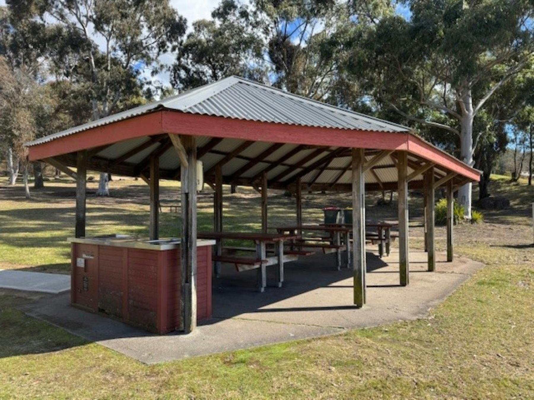 A roofed open building sheltering two BBQ's and tables and bench seating surrounded by grass