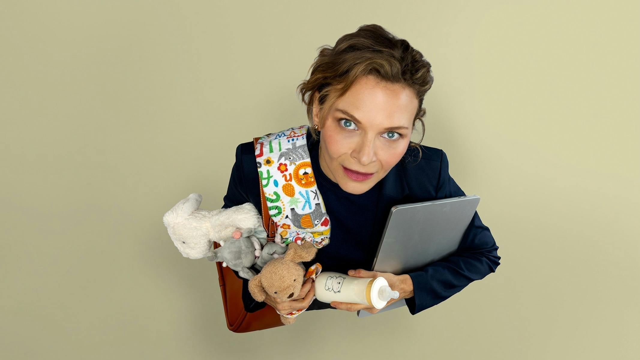 A woman stares up at the camera holding a laptop, baby blanket, plush toys and baby bottle.