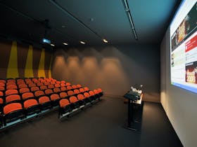A small auditorium with formal rows of orange seats, a lectern and projected image but no people
