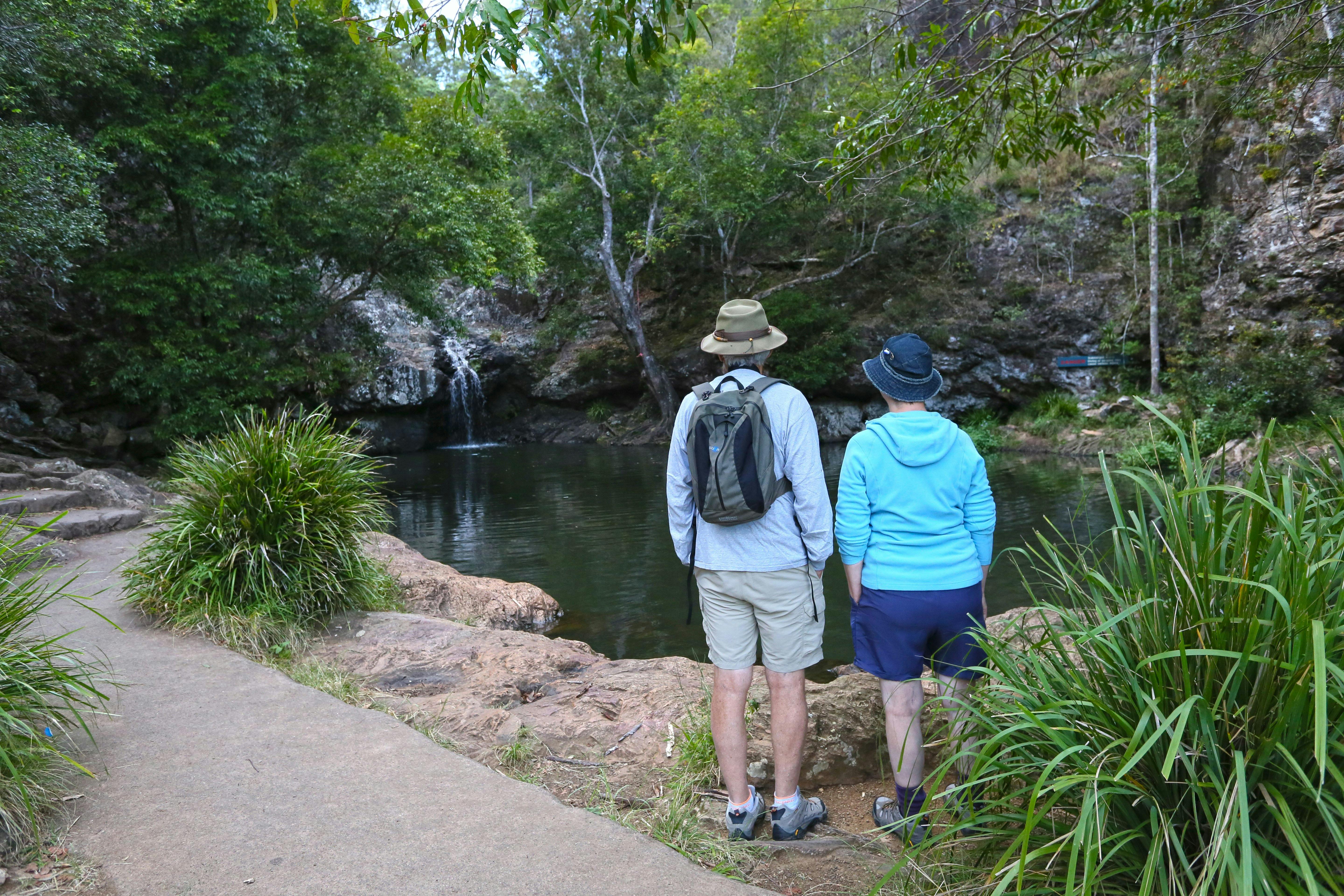 Two walkers stand gazing into a green rockpool where a waterfall cascades down a low rocky ledge.