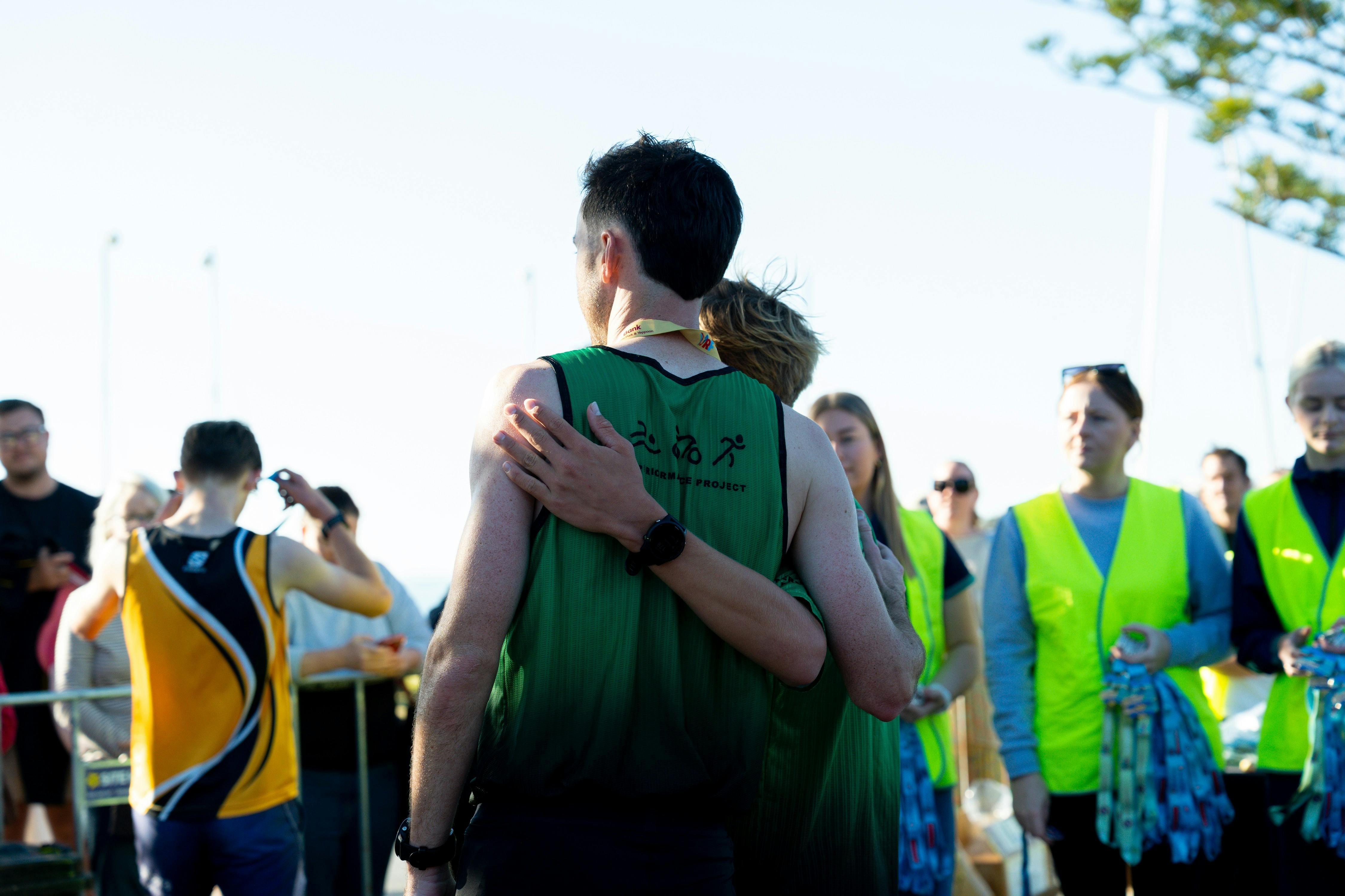 Two runners embrace in the finish area while volunteers, participants and spectators gather around t