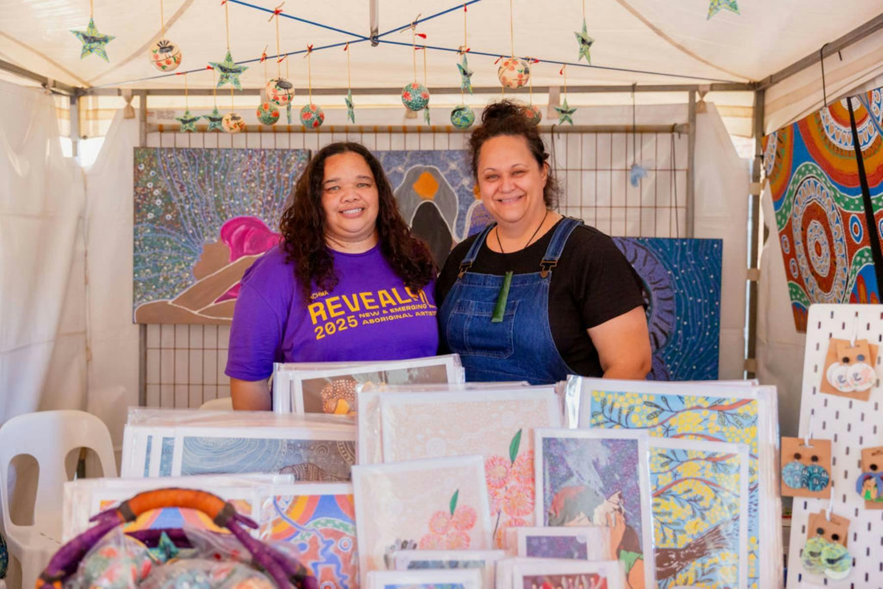 Two female Aboriginal artists stand behind their stall displaying authentic Aboriginal artworks.