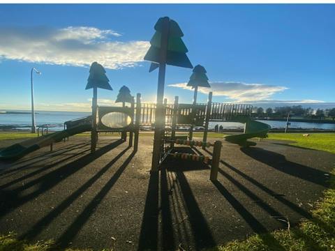 Grass area with playground equipment, overlooking the blue harbour & Norfolk pines in the distance.