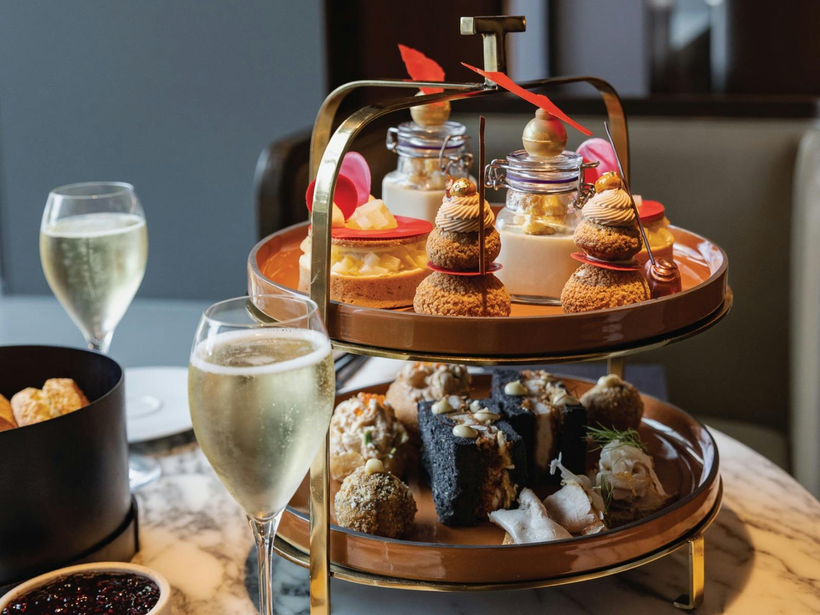 A two-tier stand of sweet and savoury high tea pastries and delicacies. Sparkling wine in foreground