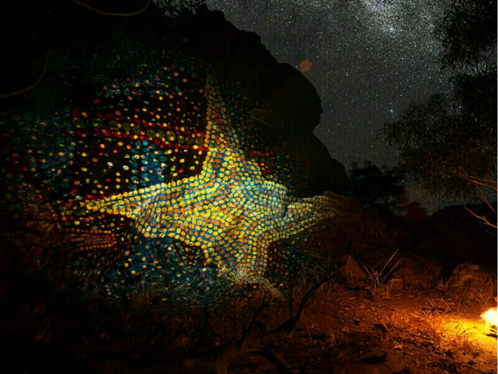 Vibrant star-shaped artwork illuminated on a rocky surface under a starlit sky, with a small fire