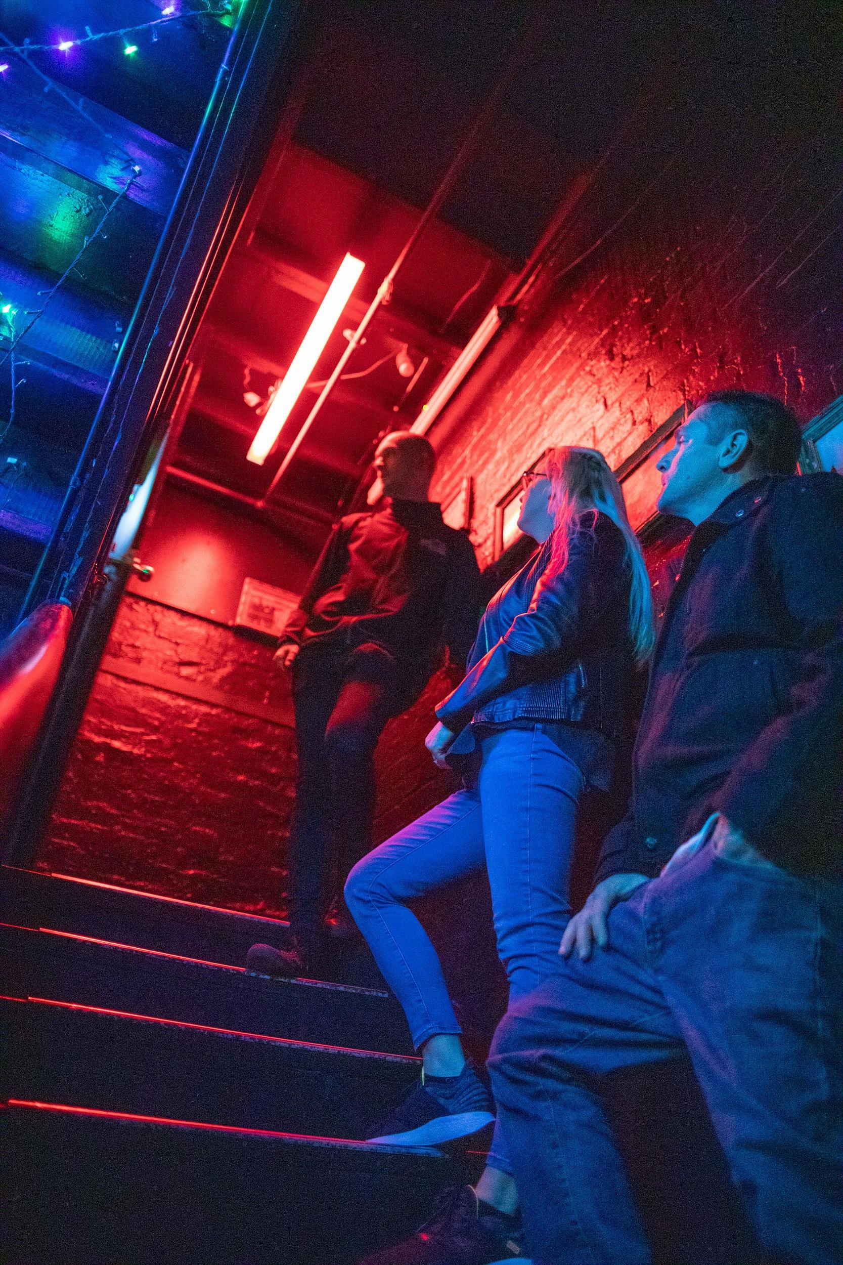 Two men and a woman standing on a dark staircase with red and blue lighting.