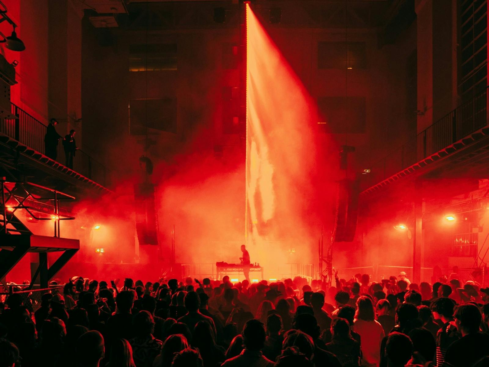 DJ performs on stage under a bright red spotlight as a large crowd fills a smoky indoor venue.