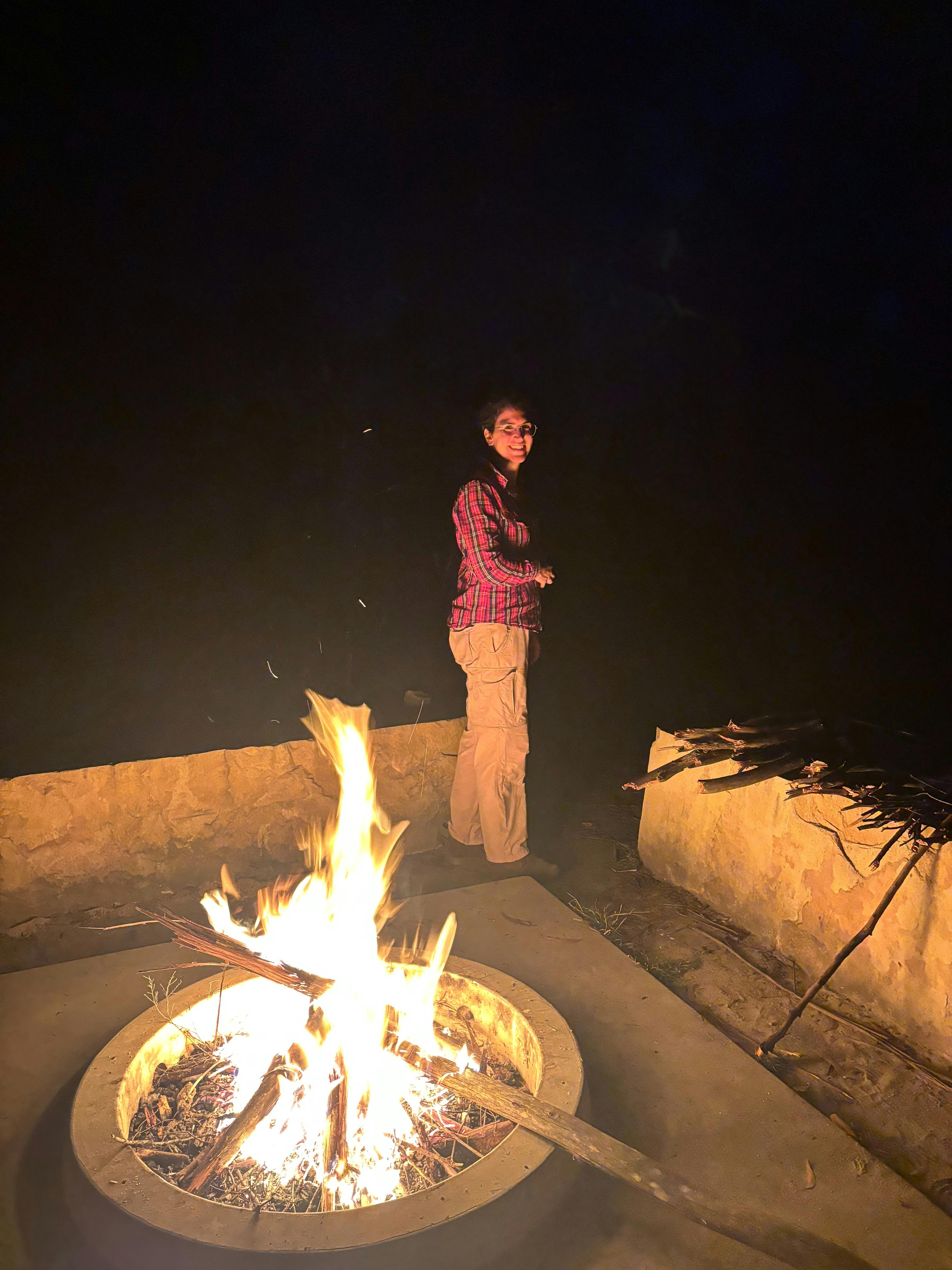 A woman wearing a red checkered shirt front lit by an inviting camp fire at night.