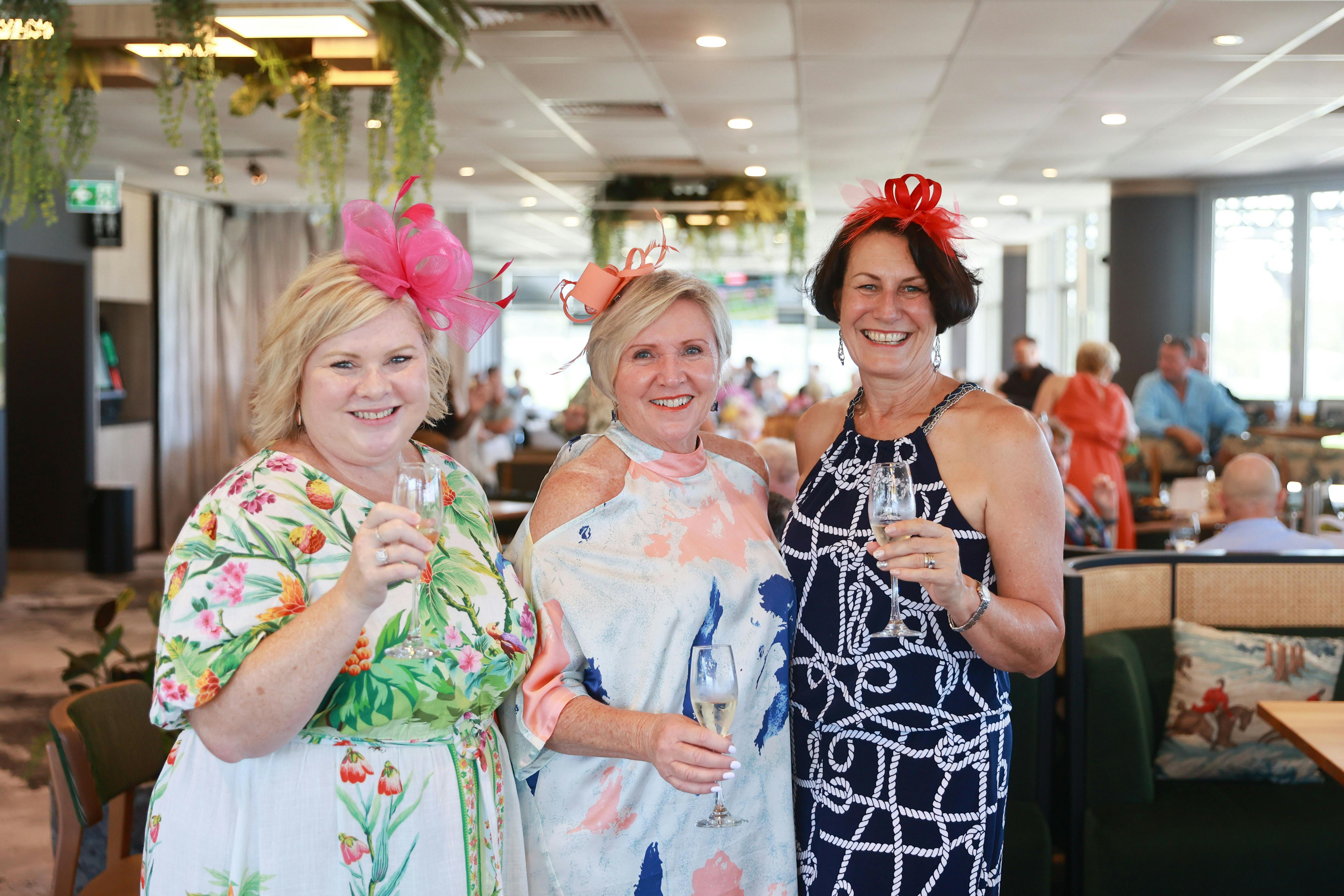 Three ladies holding drinks and smiling at the camera dressed in racewear with fascinators