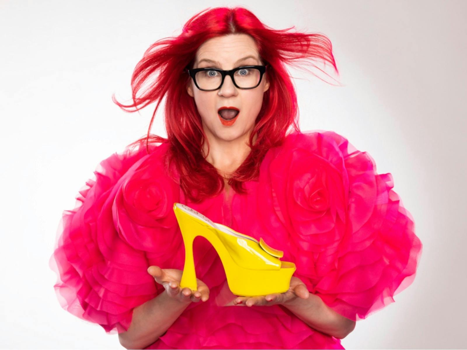 A red-haired woman in a bright pink dress holds a yellow shoe