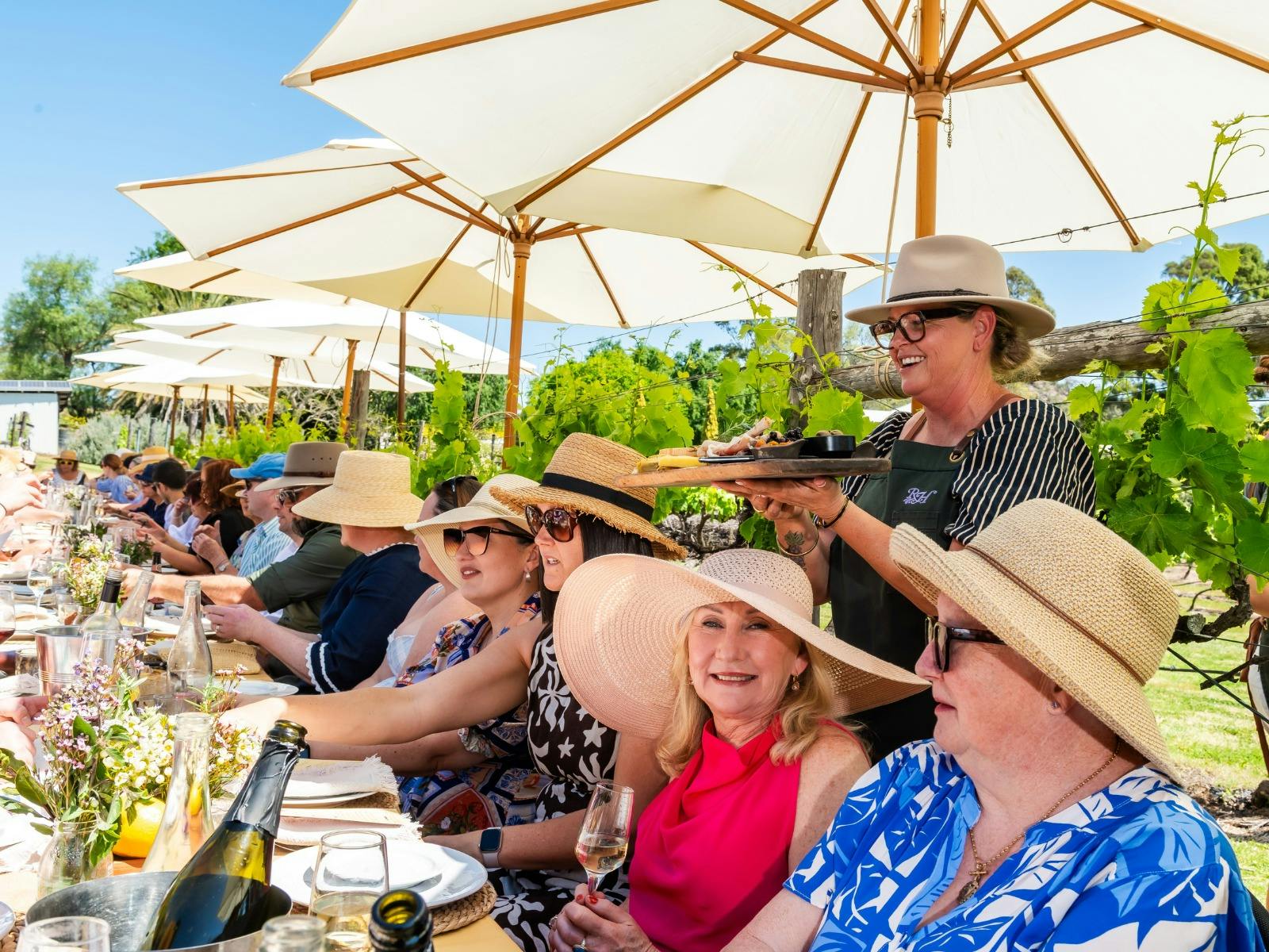 People sit at a long table under white umbrellas, enjoying a long lunch in a vineyard.