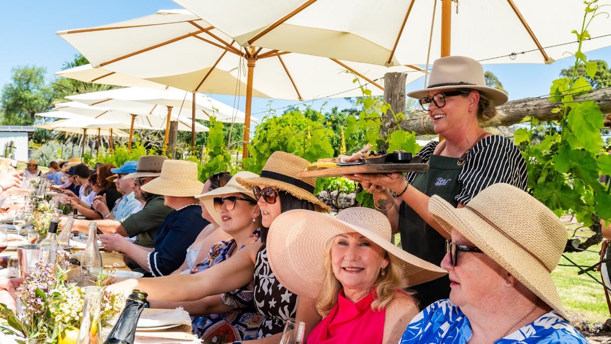 People sit at a long table under white umbrellas, enjoying a long lunch in a vineyard.