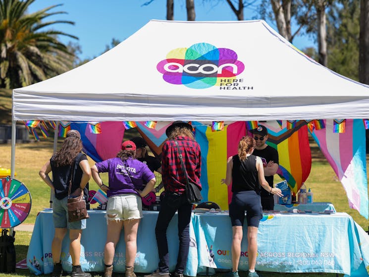 The image shows 4 people at a stall for ACON, with lots of pride flags and bright colours