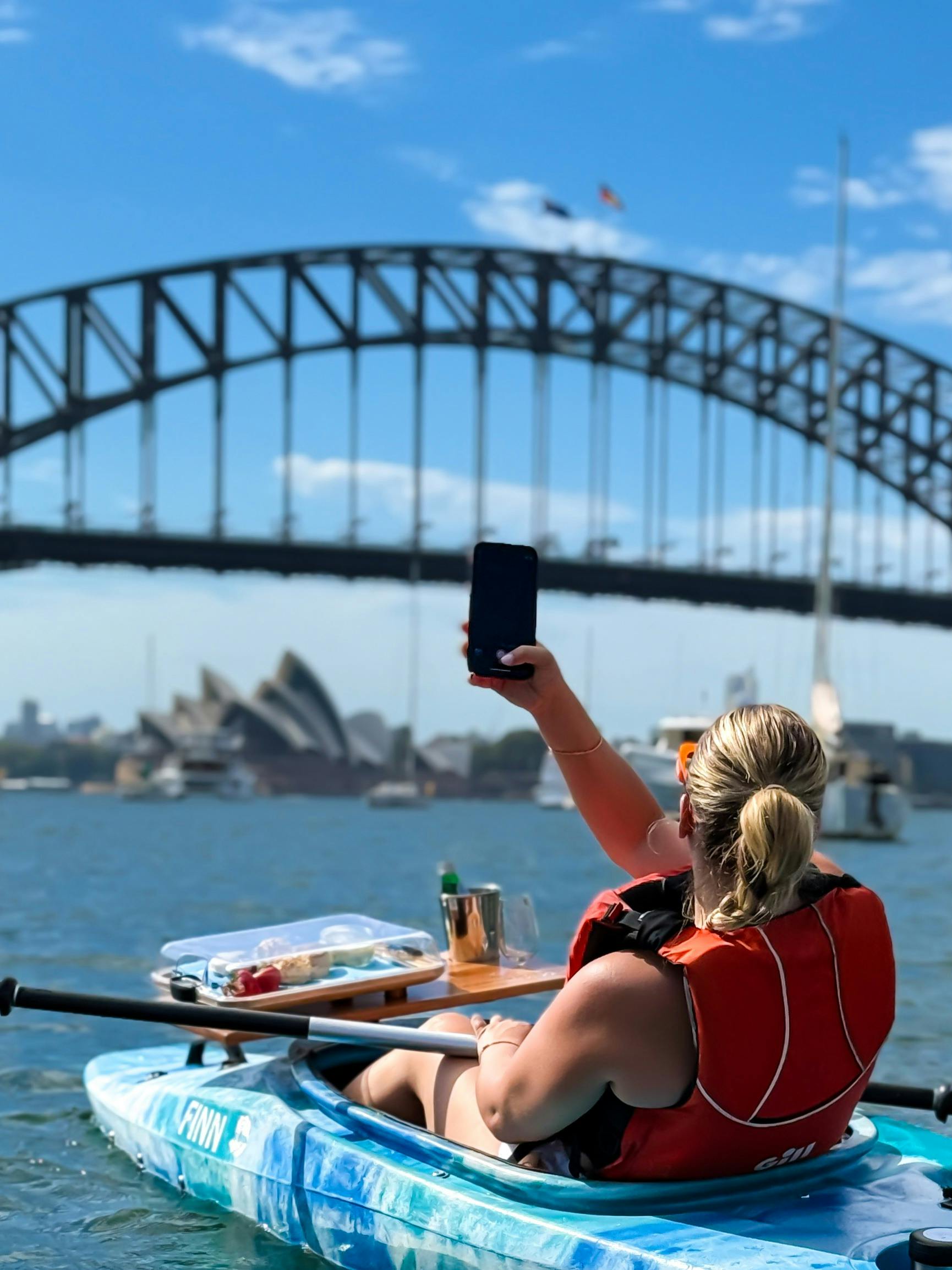 Blonde lady taking photo of Sydney Harbour Bridge and Sydney Opera House from her kayak.