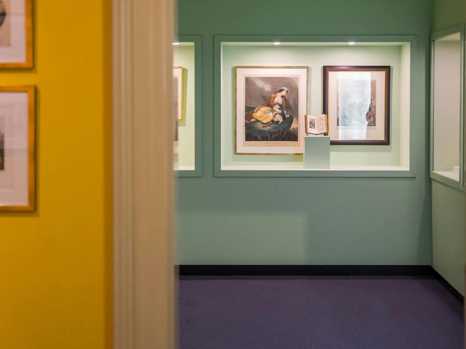 Camera looks through a doorway to a green gallery with assorted artworks, yellow wall in foreground