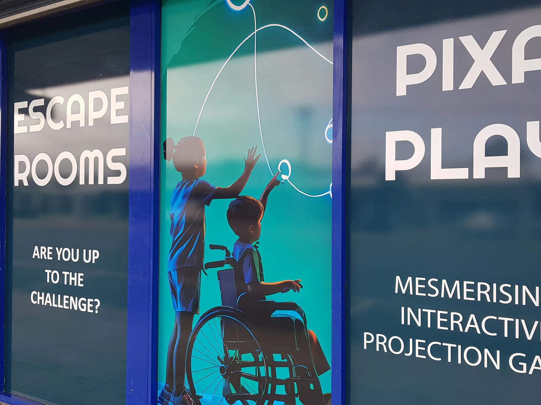 Single-level storefront with full-height signs showing people playing large-format digital games.