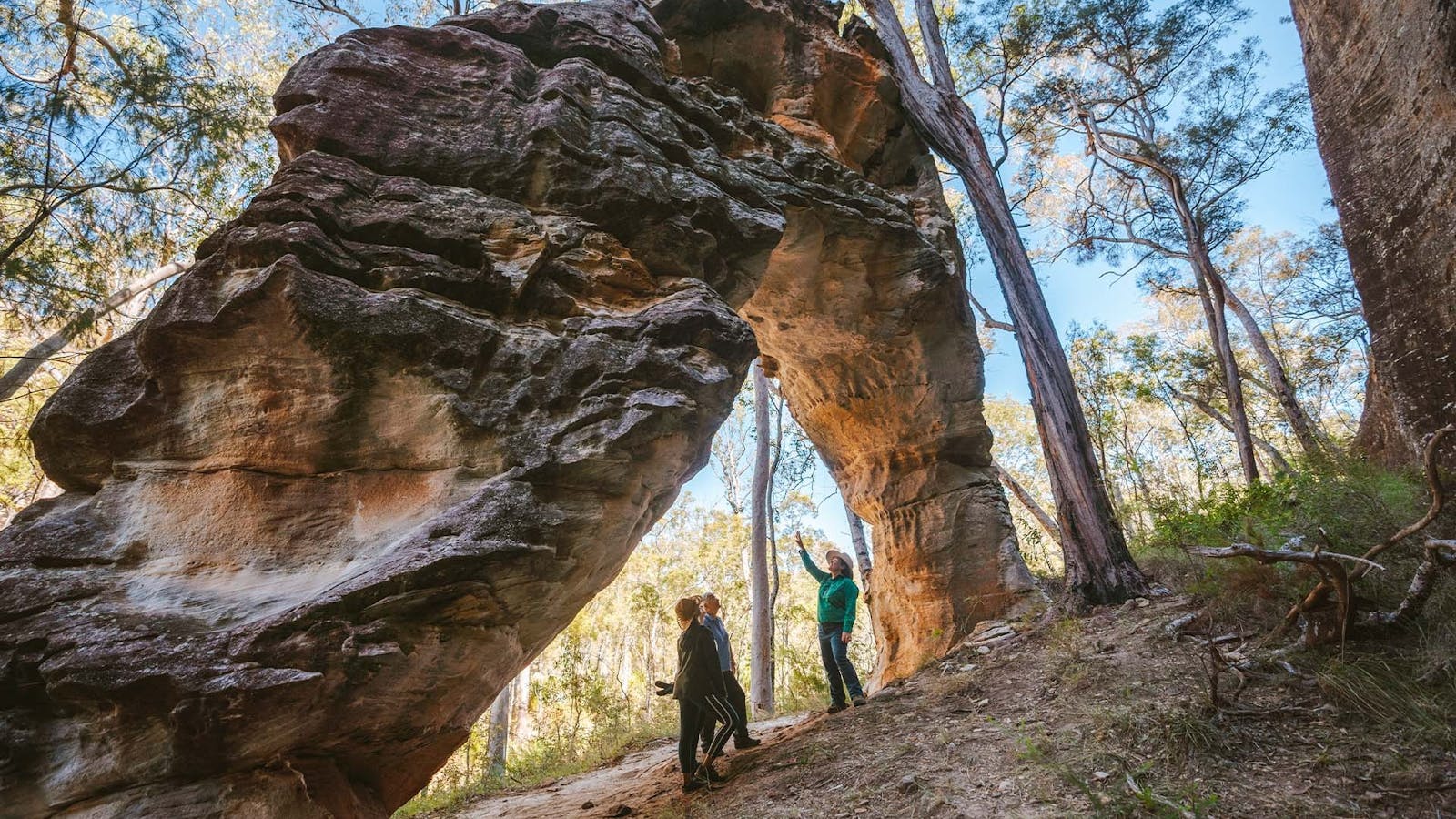 Three people standing underneath a rock arch surrounded by trees Three people standing underneath a rock arch surrounded by trees