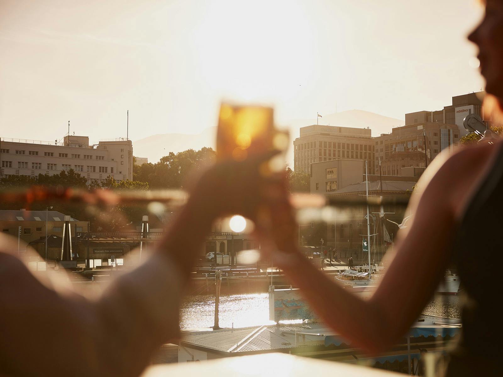 Two patrons sit on a sunny outside balcony, cheersing drinks, with a view of Hobart beyond.