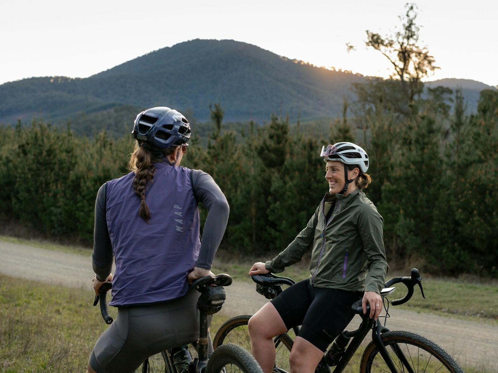 Two cyclists taking a break from riding on a gravel road with bushland and hills surrounding them.