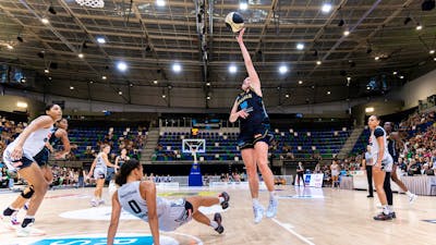A UC Capitals player shooting a basketball towards the basket while the defender slides on the floor