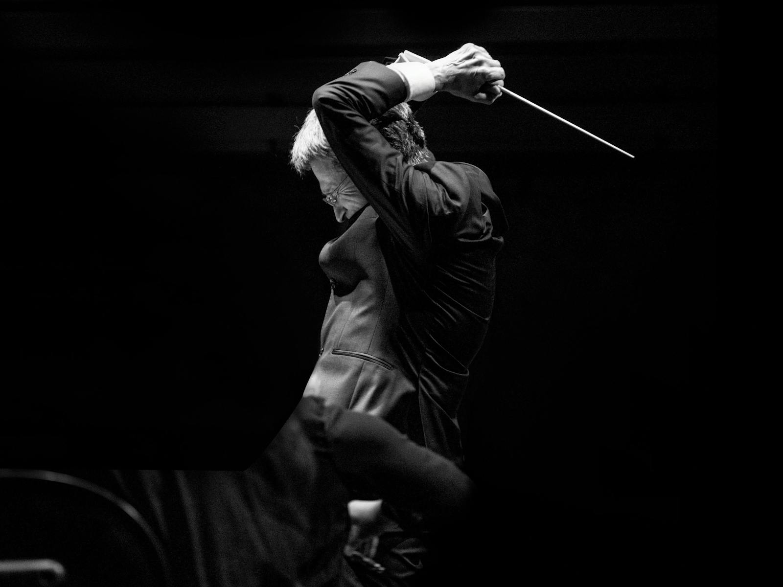 Monochrome photo of a conductor against a black backdrop. His baton is lifted behind his head.