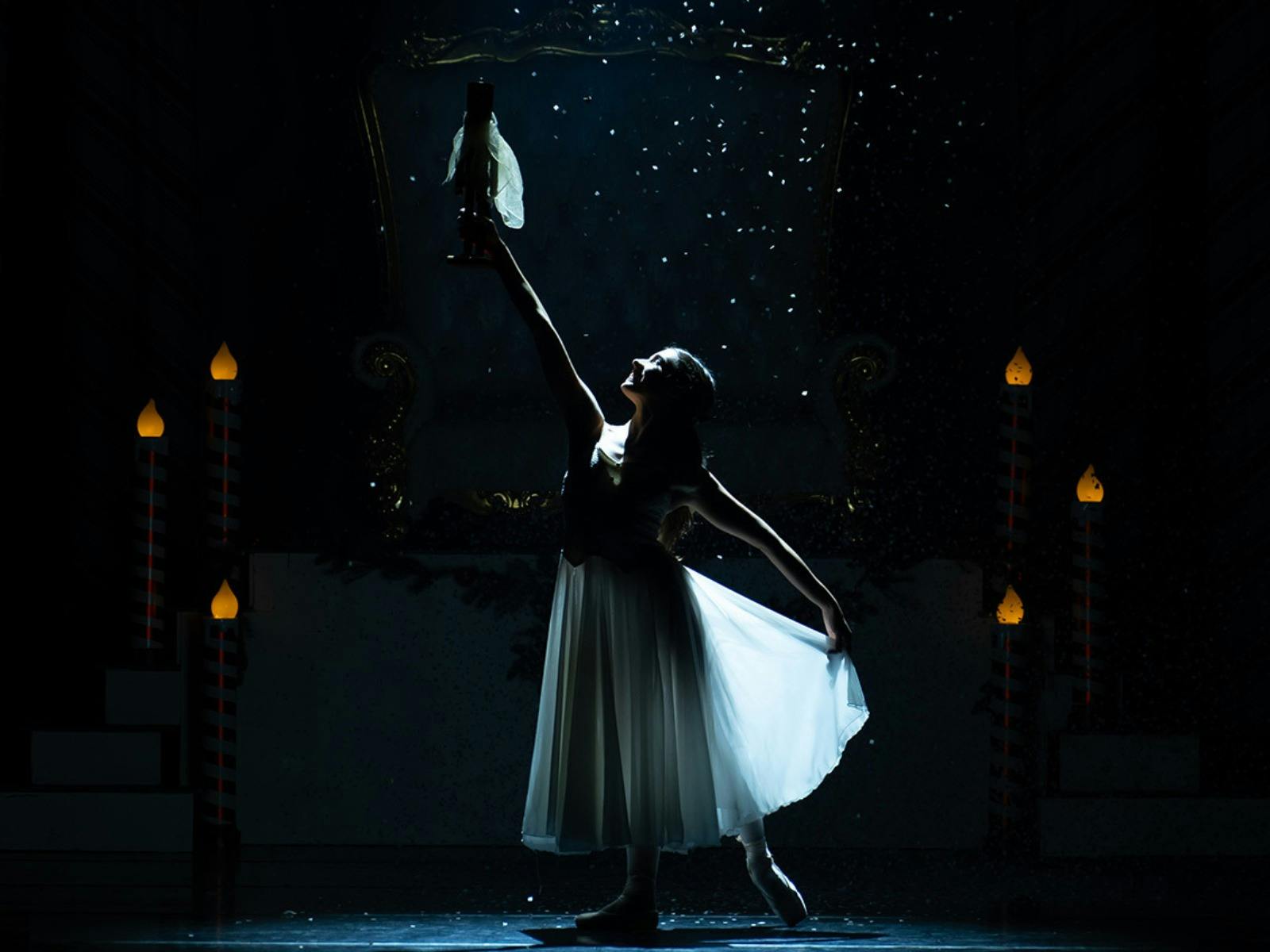 A ballerina in a white dress holds a nutcracker doll aloft under a spotlight on a dark stage.