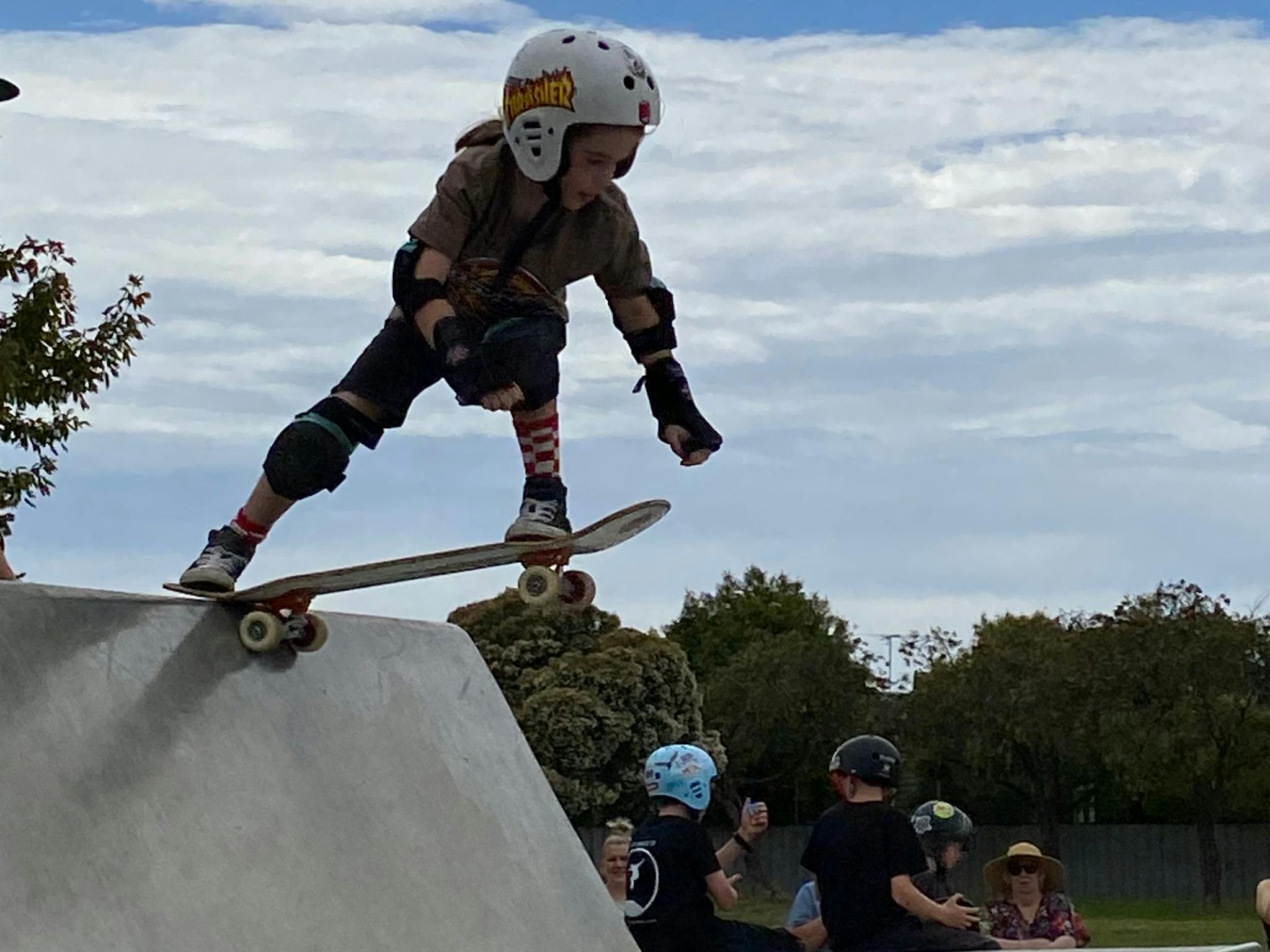Child on top of cement skate park doing a trick in the air with other people watching in background