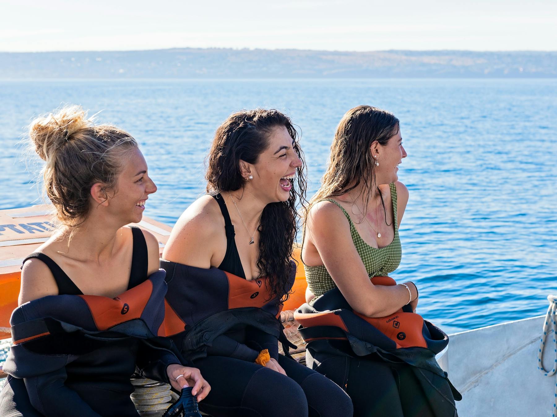 Three women wearing wetsuits that are half undone smiling sitting on the side of the Temptation boat