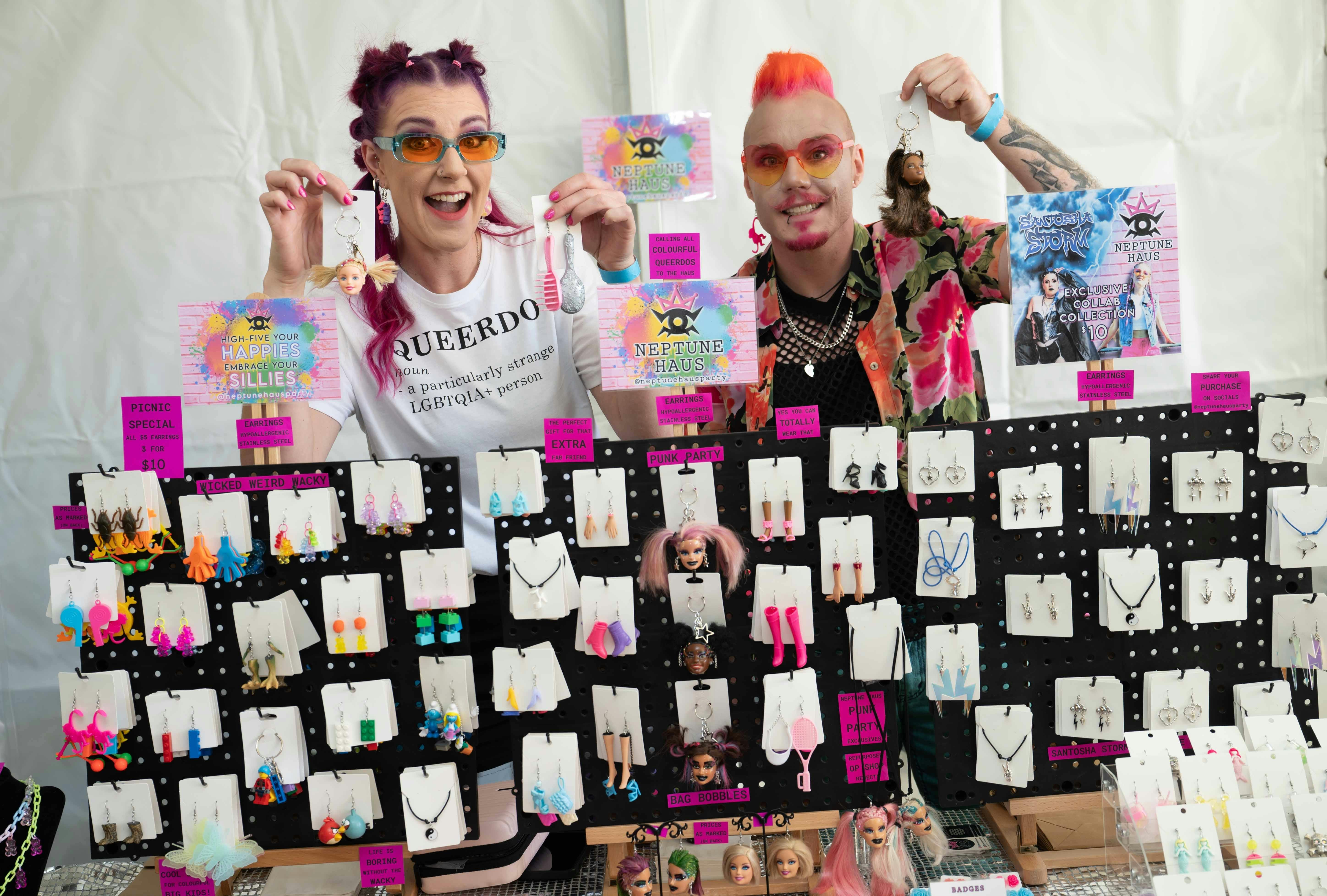 Two pink haired people holding earrings with doll heads and tiny brushes.