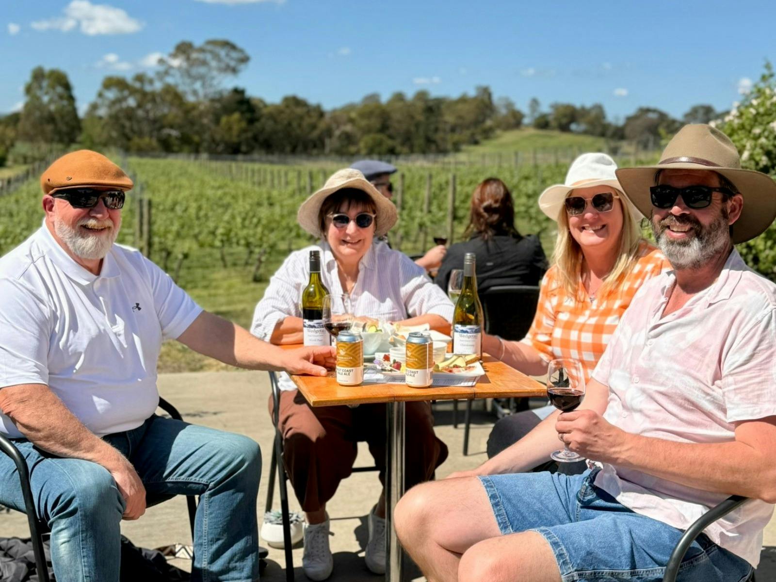 Four adults sitting at a table enjoying wine and beers with a vineyard in the background