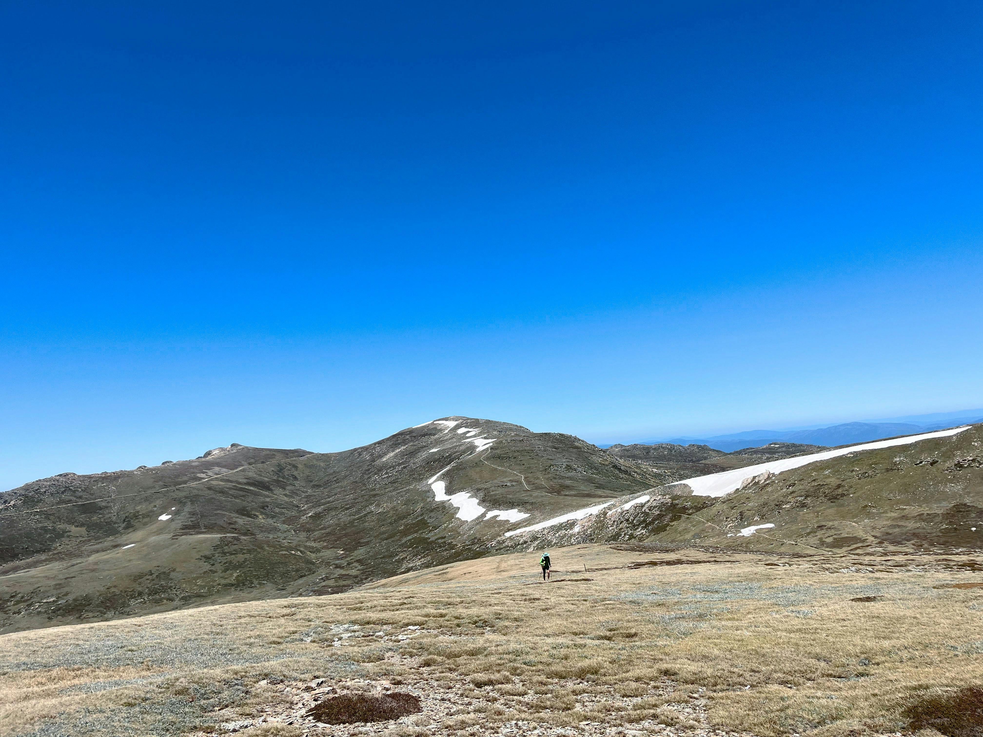 A hiker in the mountains climbing up a steady hill with snow in the background.