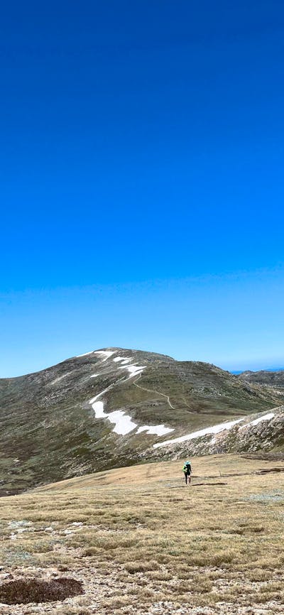 A hiker in the mountains climbing up a steady hill with snow in the background.