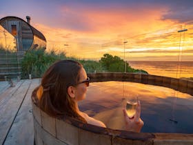 woman relaxing in timber hot tub with glass of champagne while looking at ocean