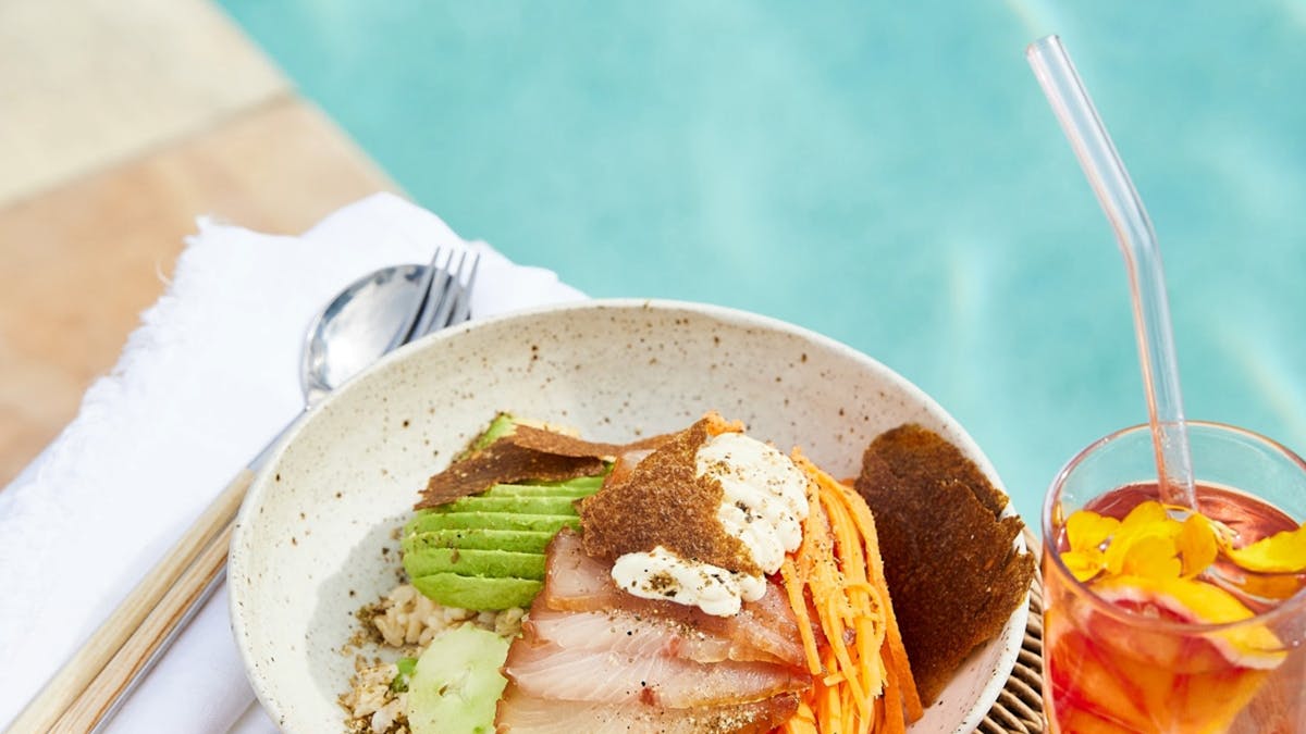 Bowl of colourful vegetables and fish with an iced tea overlooking pool