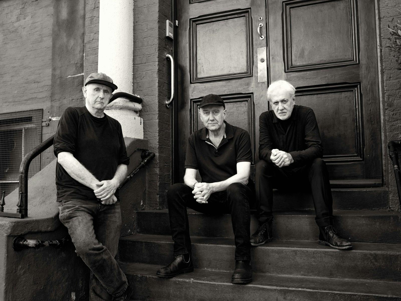 Three older men in dark clothing and hats sit and lean on the steps of a brick building