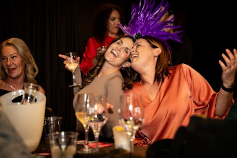 Two ladies sitting with heads together are singing happily holding glasses of wine