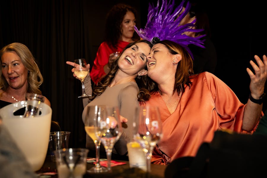 Two ladies sitting with heads together are singing happily holding glasses of wine