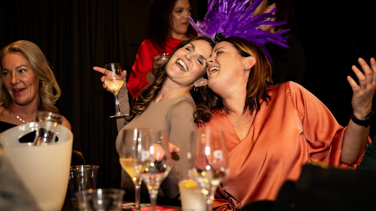 Two ladies sitting with heads together are singing happily holding glasses of wine