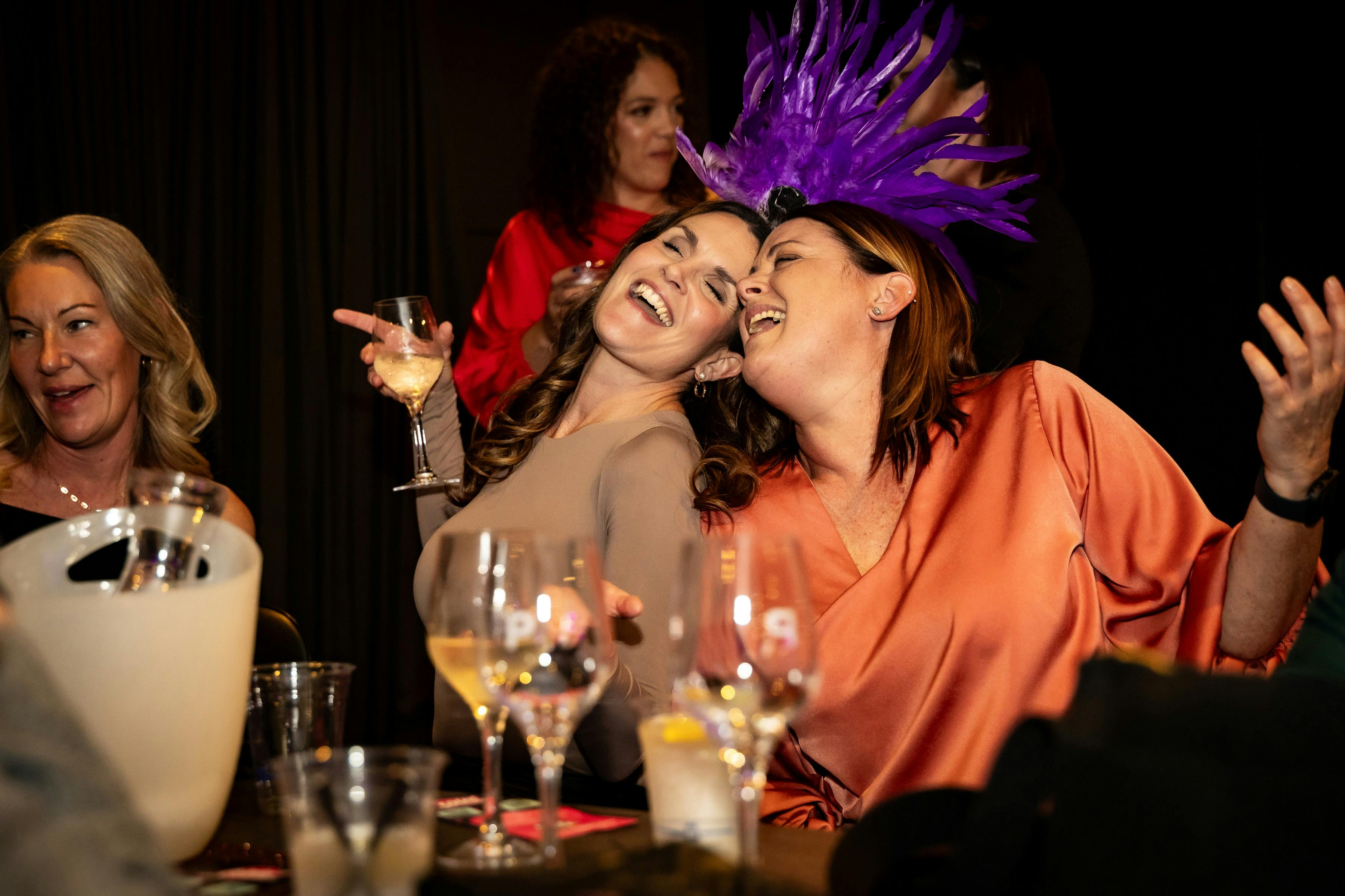 Two ladies sitting with heads together are singing happily holding glasses of wine