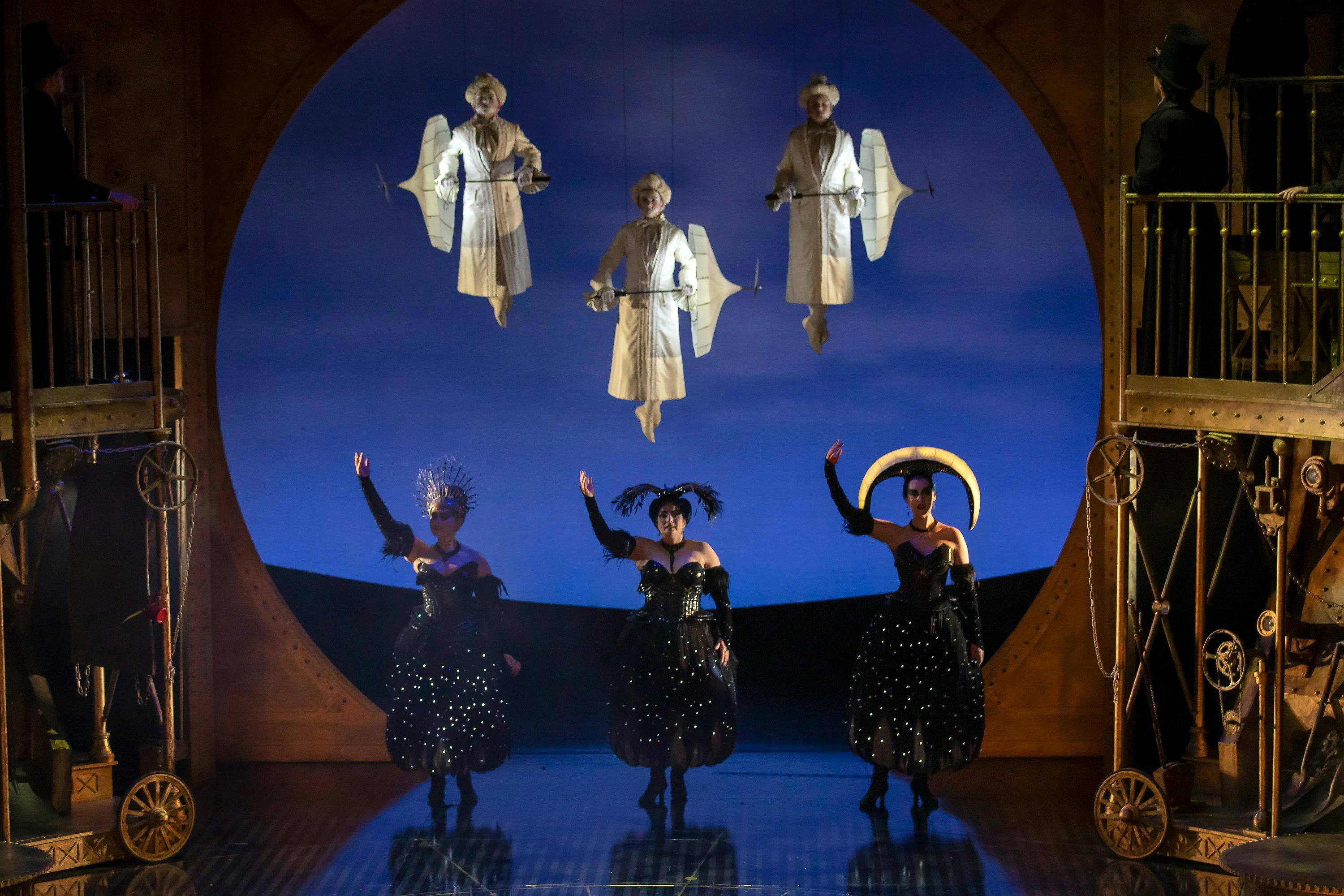 Black-clad performers pose below three floating figures in white costumes on a blue-lit stage.
