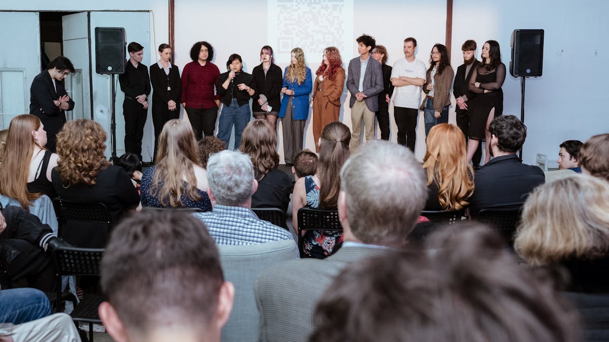 A group of people stand in a line facing an audience. One person speaks into a microphone.