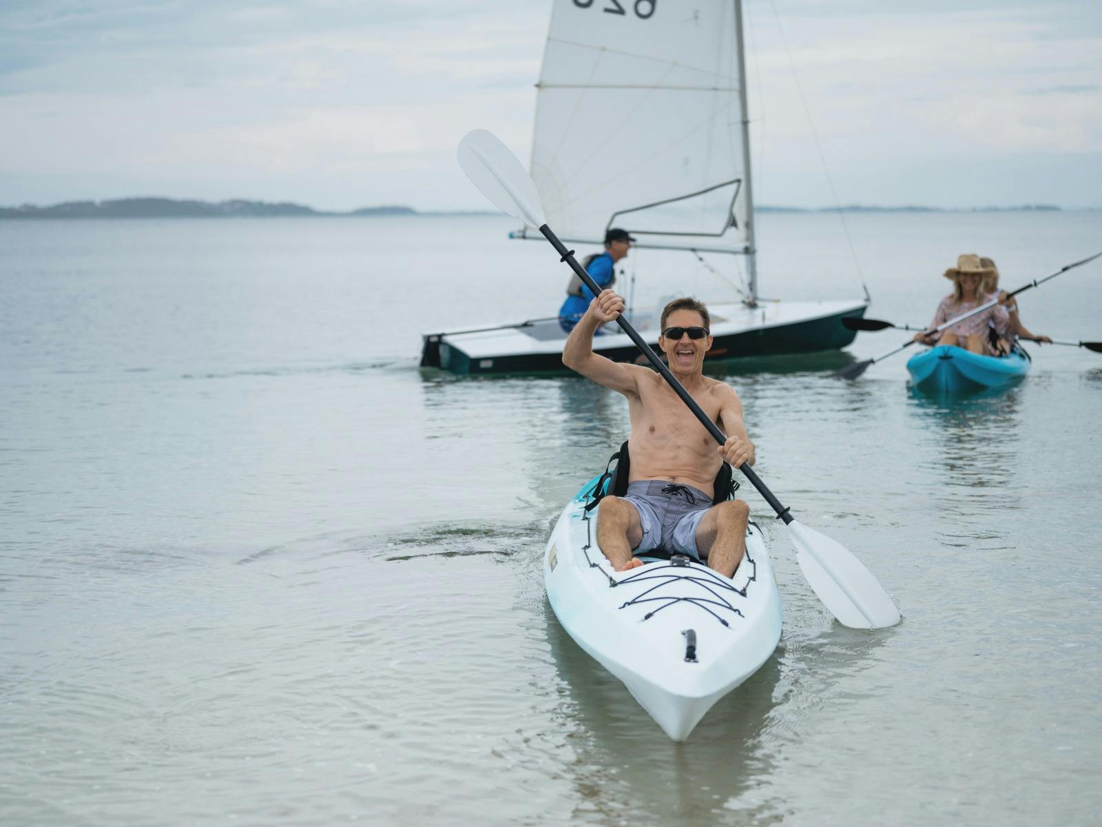 Man smiling from his kayak, two women kayaking together behind him and a sail boat in the background