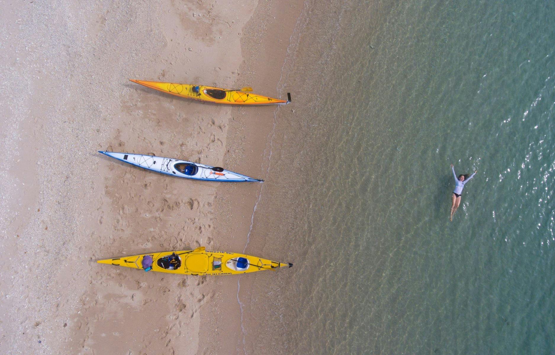 A person floats in crystal clear waters in front of a sandy beach upon which 3 kayaks rest