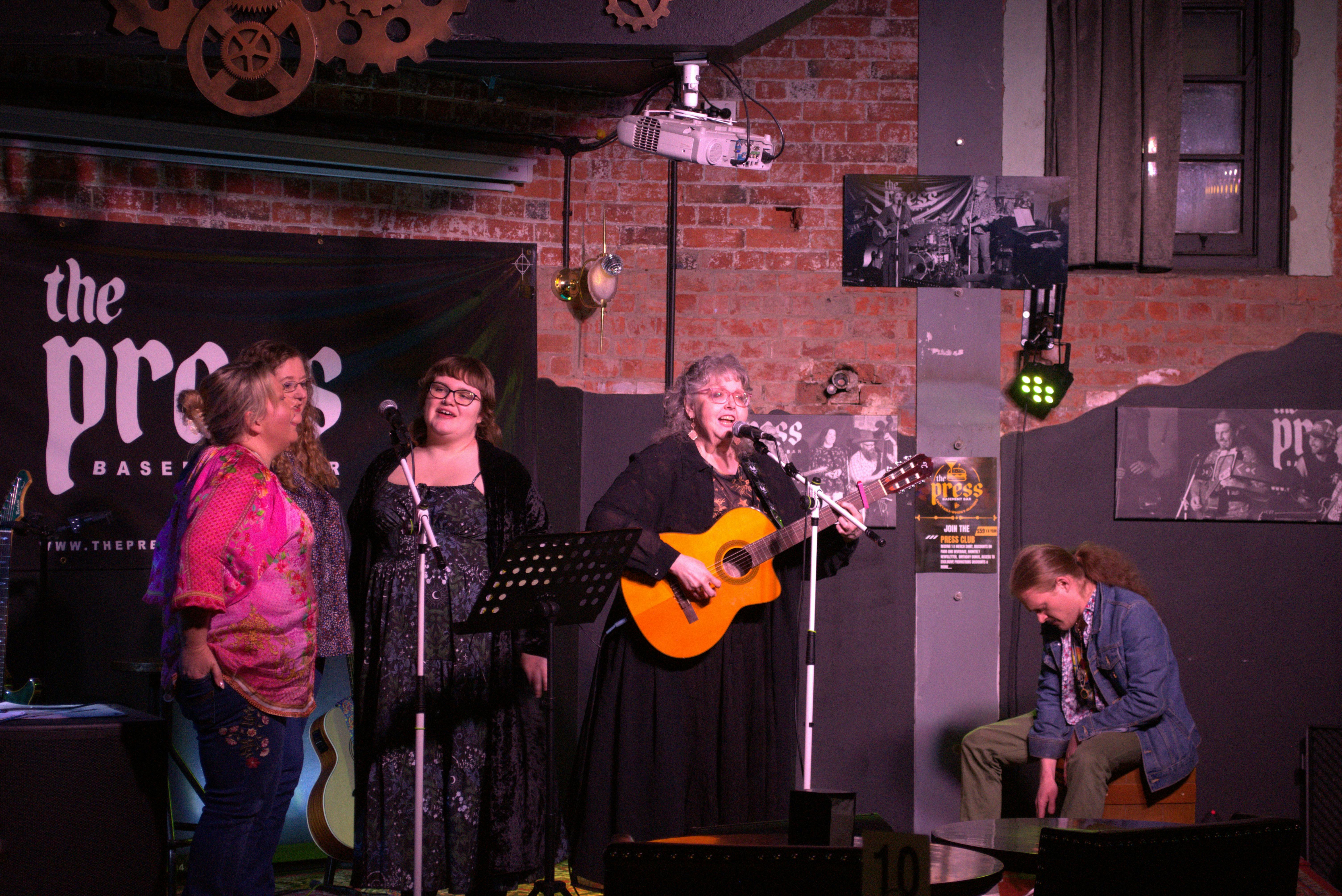 Lady in black dress plays guitar at the Press Basement Bar. 3 backing singers and a cajon player