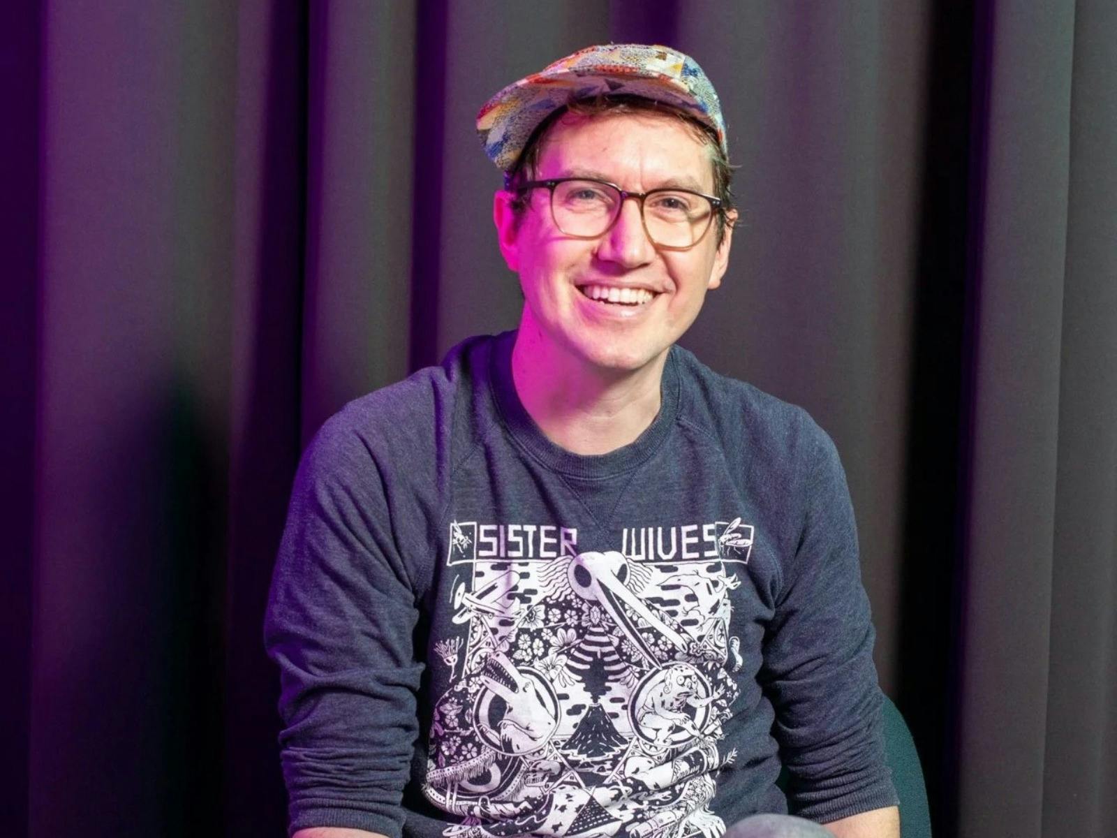 A smiling man wearing glasses and a colorful cap, seated in front of a dark curtain.